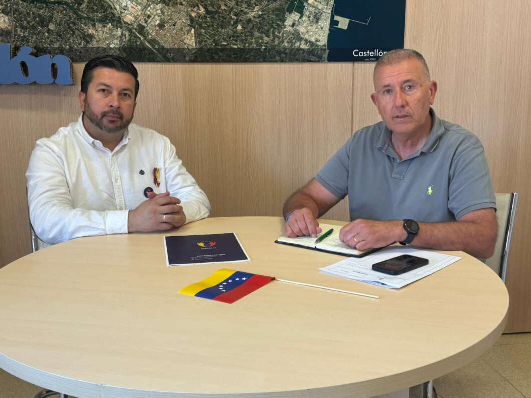 Two men sit at a round table in an office, with documents and a small Venezuelan flag on the table as they look at the camera.