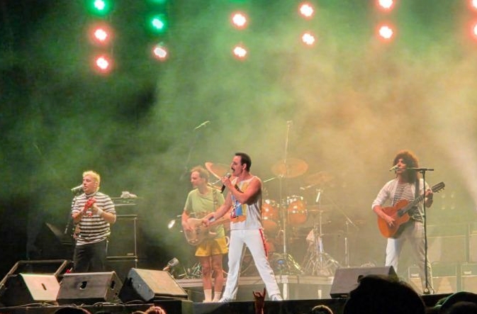 Five musicians perform on a lit stage: lead singer center, guitarist left, bassist behind, drummer in back; colorful stage lights and fog.