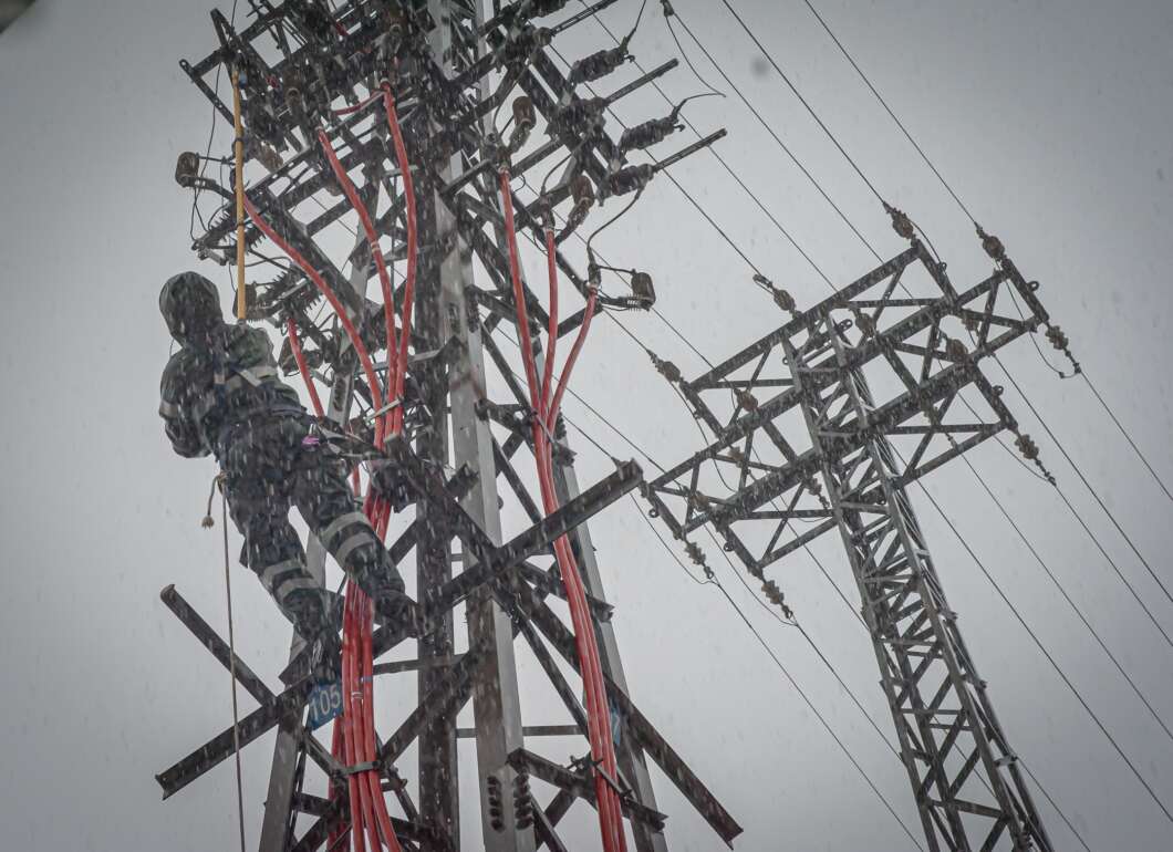 Lineman in protective gear climbs a metal utility pole amid snow and tangled red cables and power lines.