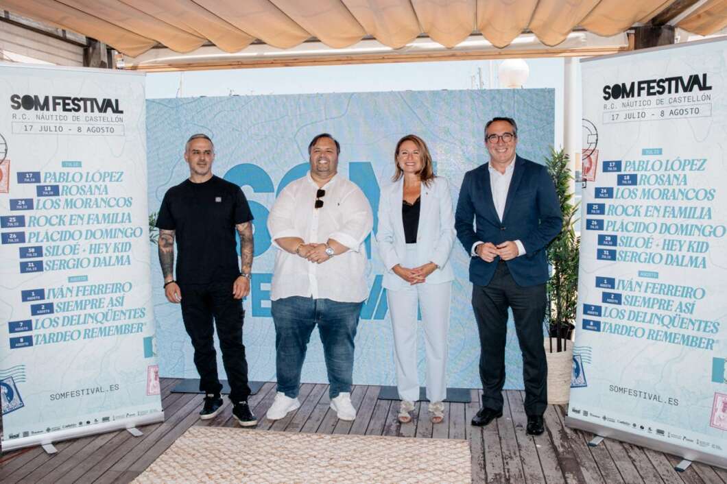 Four people stand on a wooden stage between two SOM Festival banners, posing for a photo in smart-casual to formal attire.