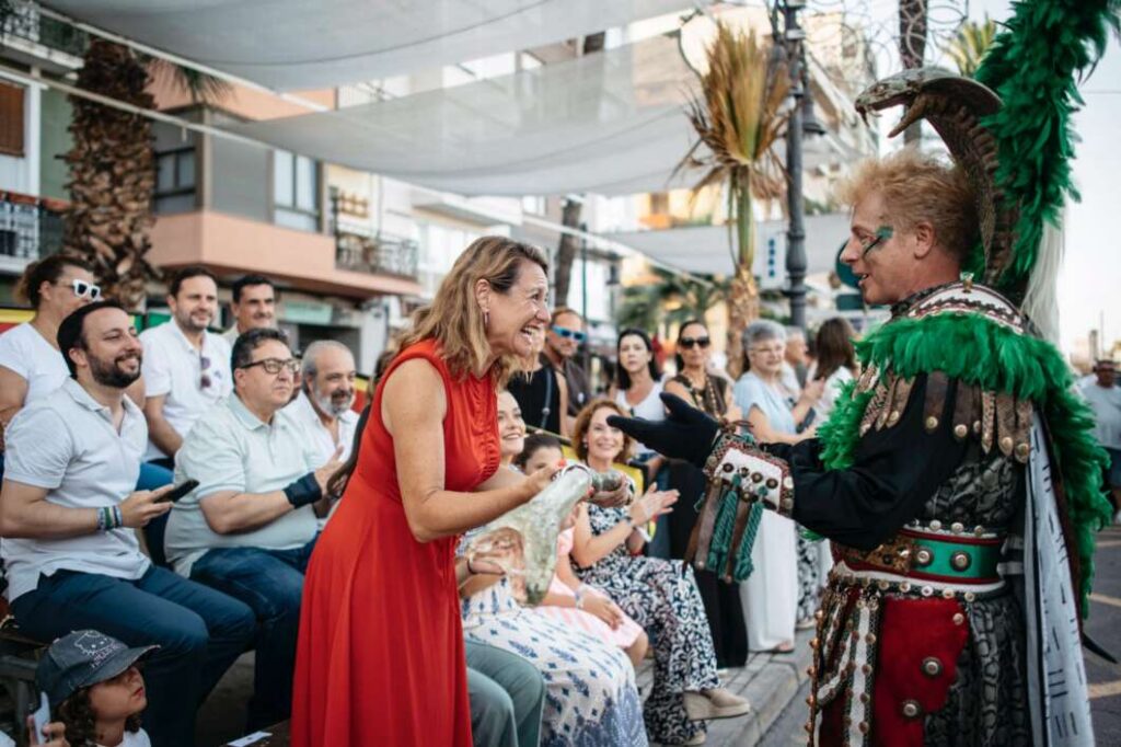 Audience at a sunny street festival watches a costumed performer with green feathers greet a smiling woman in a red dress who hands him a bottle.