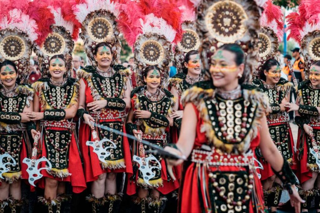 Group of dancers in red and black traditional costumes with large circular feather headdresses at a parade, smiling and posing in formation.