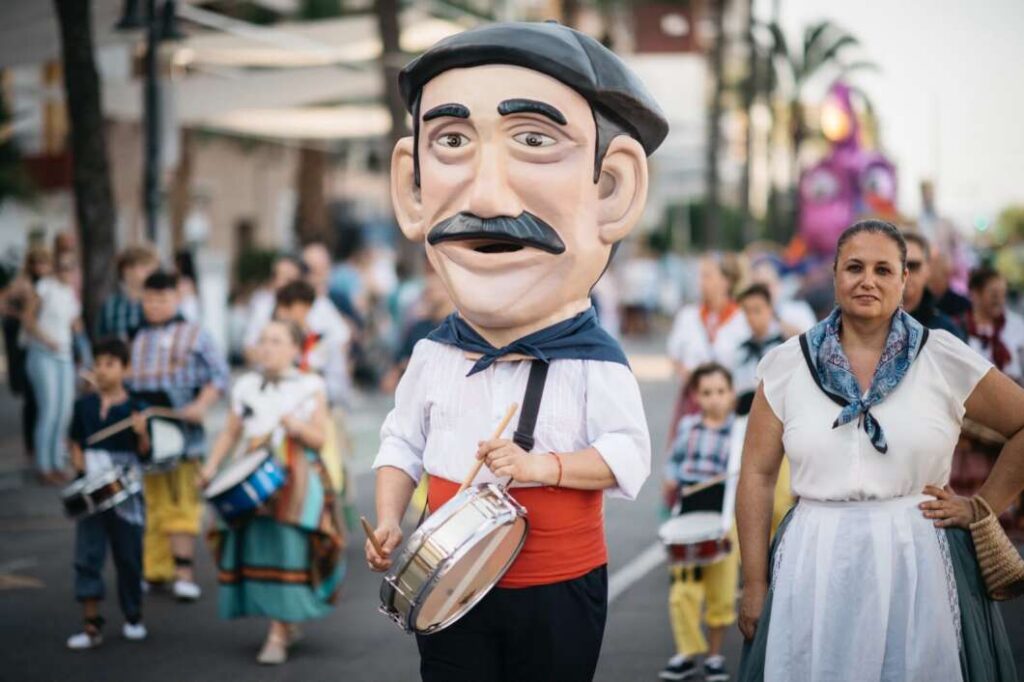 Person in a large mascot head costume playing a snare drum at a street parade, with spectators around. (Informative)
