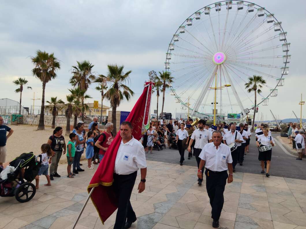 Parade on a beach promenade with a large Ferris wheel in the background; flag bearers march ahead while spectators line the walkway.