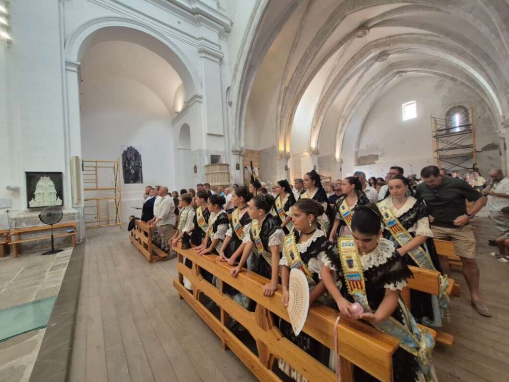 Group of girls in traditional dresses and sashes kneeling in wooden pews inside a vaulted church.