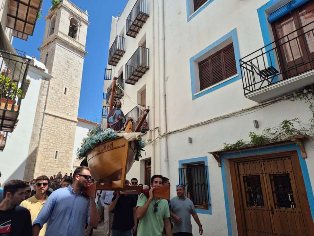 Religious procession on a narrow sunlit street, people carry a wooden boat-shaped float with a saint statue adorned with flowers in front of white buildings with blue trim.
