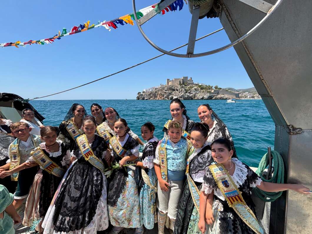 Group of girls in colorful traditional dresses wearing sashes on a boat, with blue sea and a fortress island in the background.