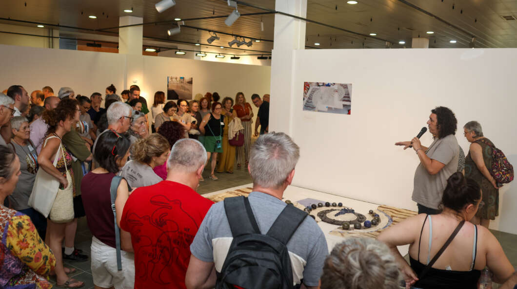 Speaker with microphone addresses a crowd at an art exhibit, standing beside a circular bead installation on the floor.