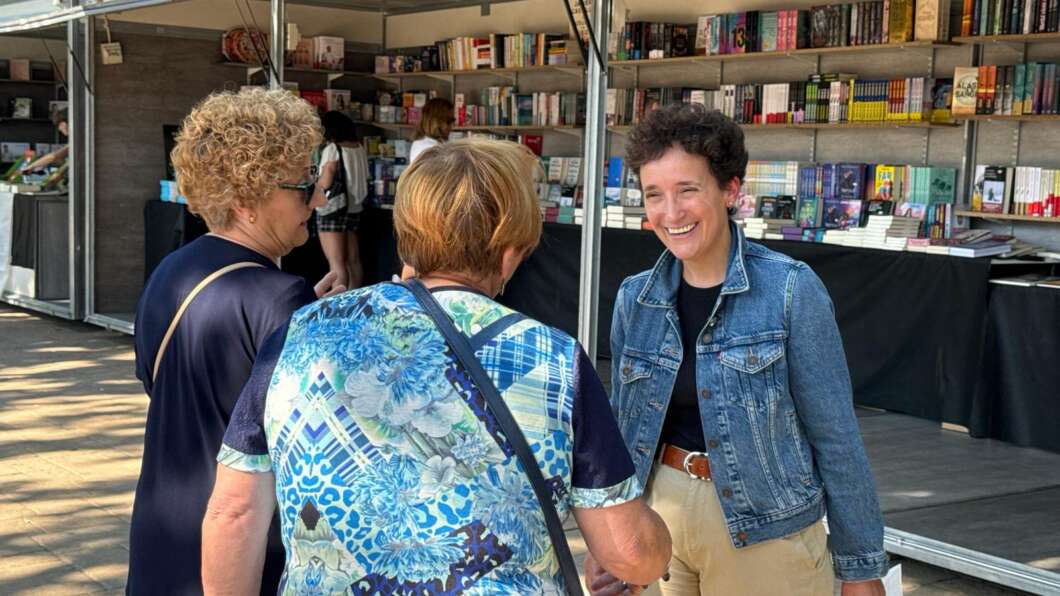 Smiling woman in a denim jacket greets two customers at an outdoor book market stall with shelves of books behind them.