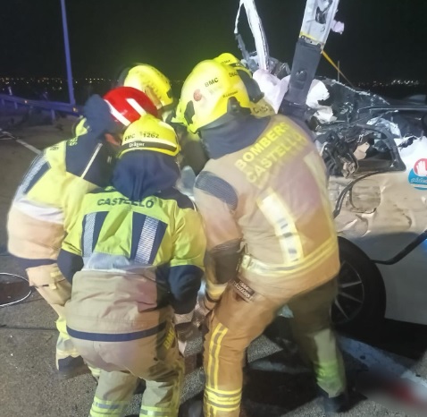 Nighttime rescue scene: firefighters in yellow turnout gear working around a damaged white car on a highway, using tools to access the vehicle