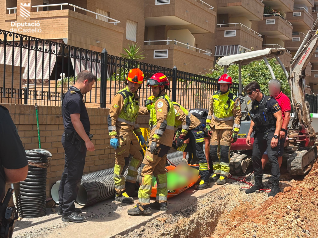 Emergency responders, including firefighters and police, gather around a blurred person on an orange stretcher in a trench next to a construction site with a backhoe and coiled hoses nearby.
