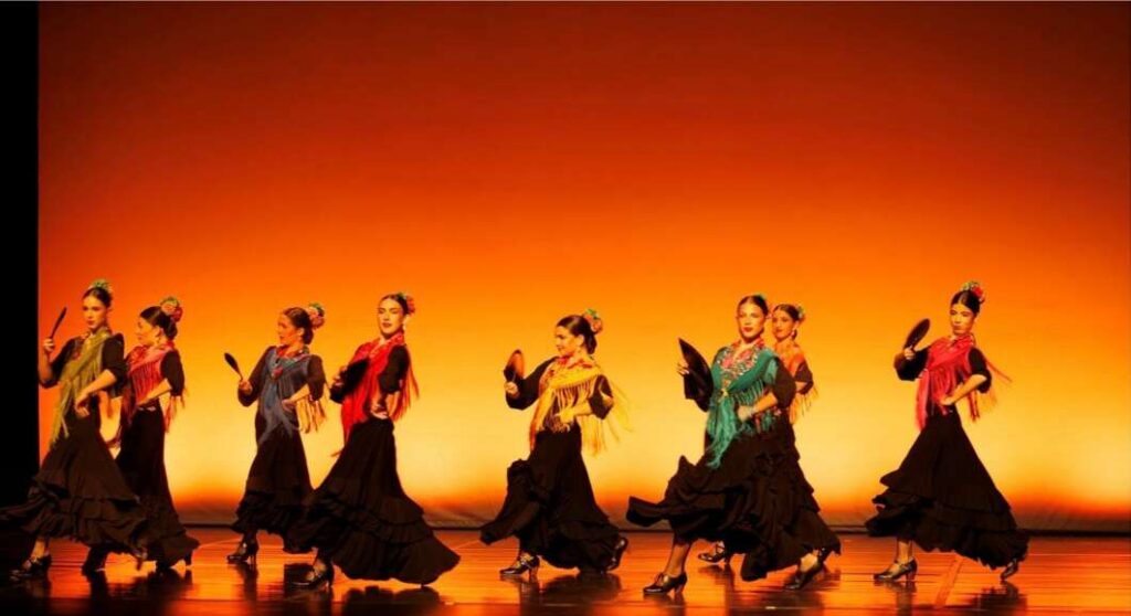 Group of female dancers in long black skirts and colorful shawls performing a fan dance on a warm orange-lit stage.