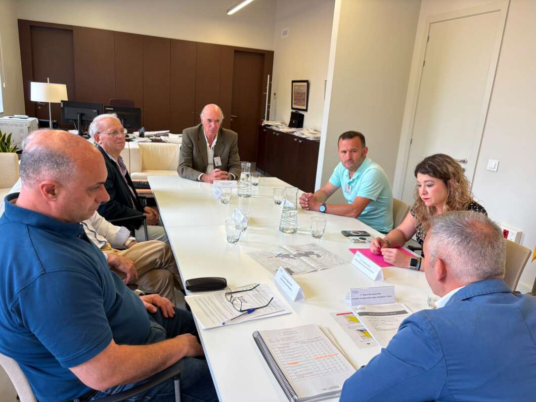 Six people sit around a long conference table in a modern office, with papers, notebooks, and water glasses on the table.