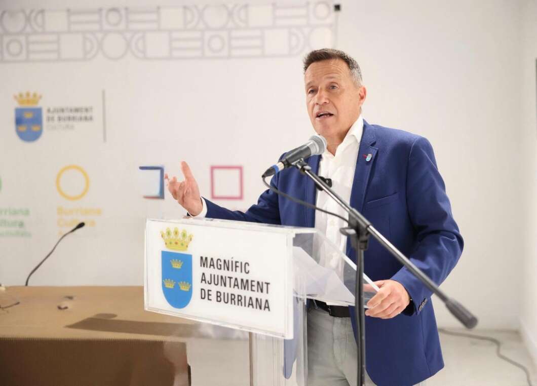 Man in a blue blazer speaks at a podium with a microphone during a municipal event; podium shows a crest and the words 'Magnífic Ajuntament de Burriana'.
