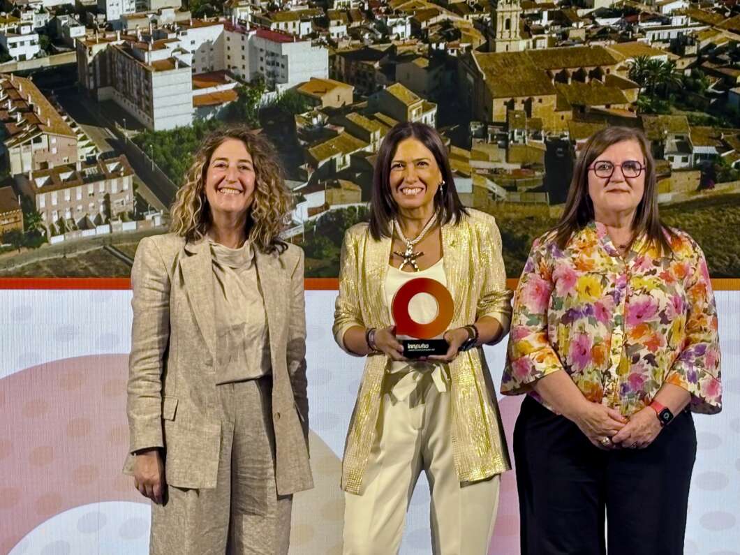 Three women stand on a stage; the center woman holds a red circular trophy against a town backdrop