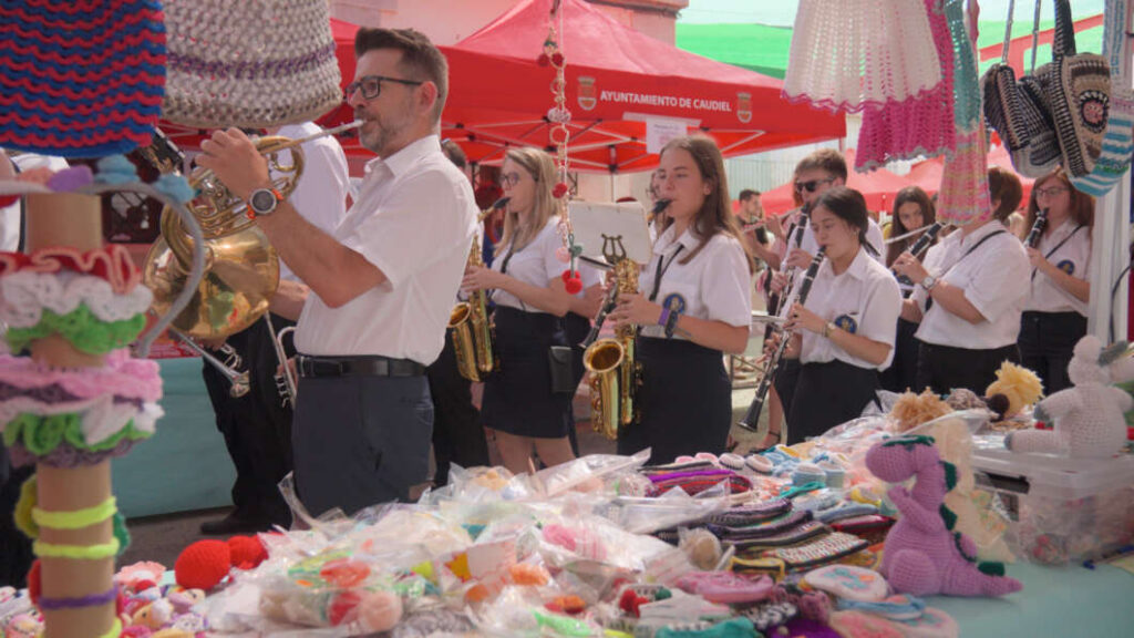 Brass band performing at an outdoor market, with colorful handmade crafts laid out on a table in the foreground.