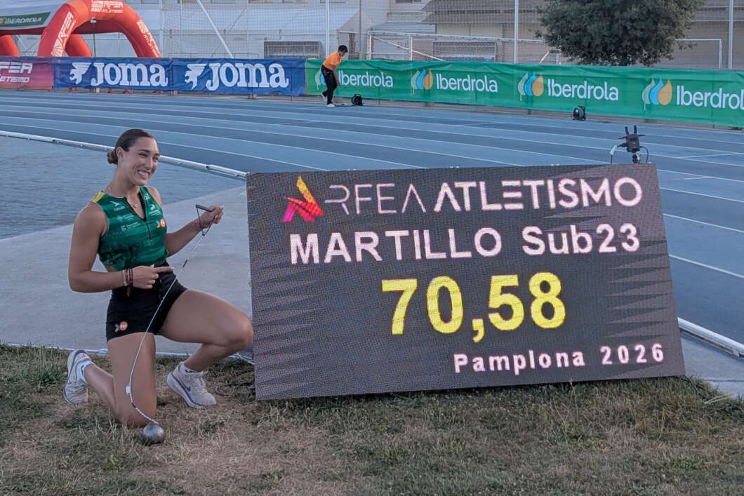 Female hammer thrower kneels beside a digital scoreboard reading REAL ATLETISMO MARTILLO SUB23 70,58 Pamplona 2026, smiling at the camera with a hammer in hand and track behind.