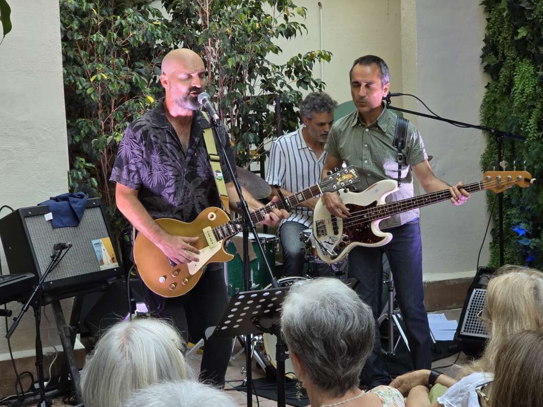 Band performing outdoors: a bald guitarist sings into a microphone while playing a gold Les Paul; a bassist and drummer accompany him; audience watches nearby.