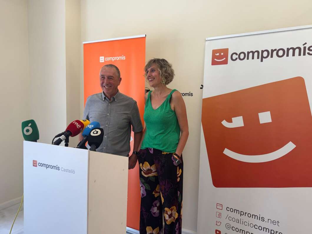 Man and woman at a podium with multiple microphones, smiling during a Compromís press event with orange banners behind them.