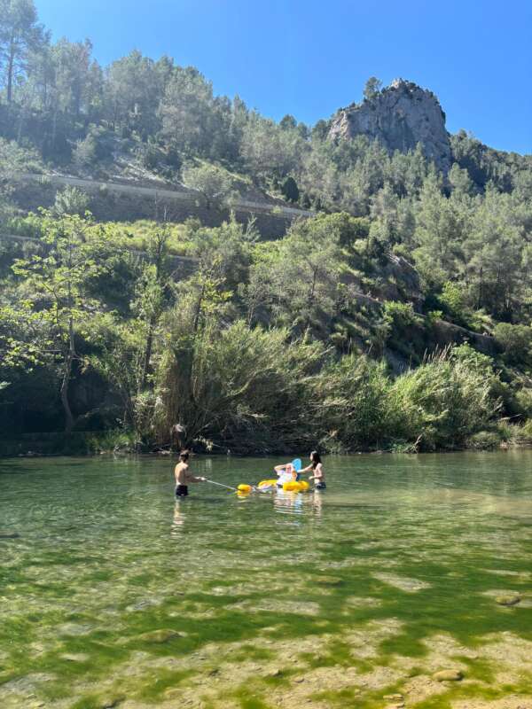 Three people in a clear green river with yellow inflatable rings, surrounded by trees and a rocky hill under a blue sky.