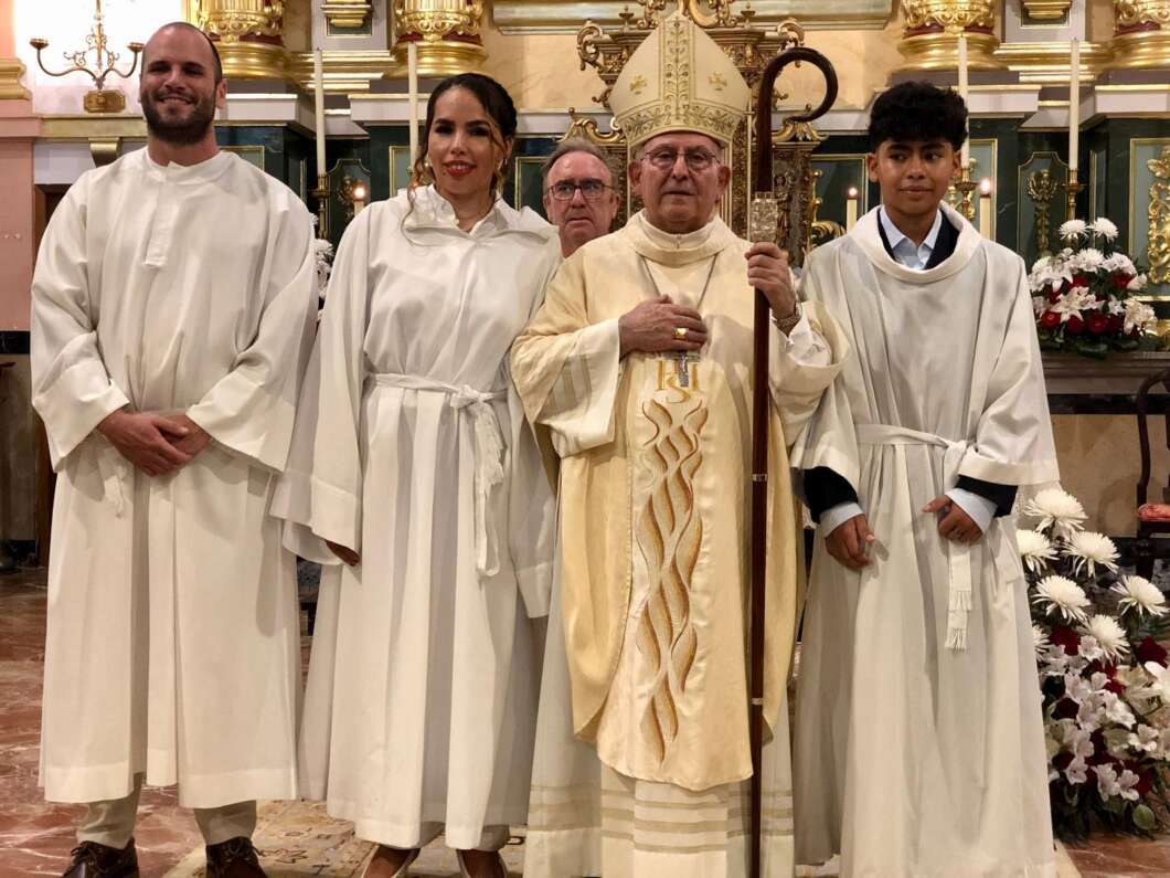 Catholic bishop in cream vestments holding a staff, flanked by four altar servers in white robes inside a decorated church.