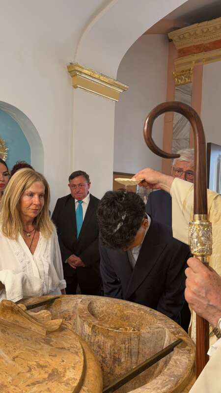 Group of people gathered around a large stone baptismal font during a religious ceremony; a priest holds a staff while a man in a suit bows his head near the water.