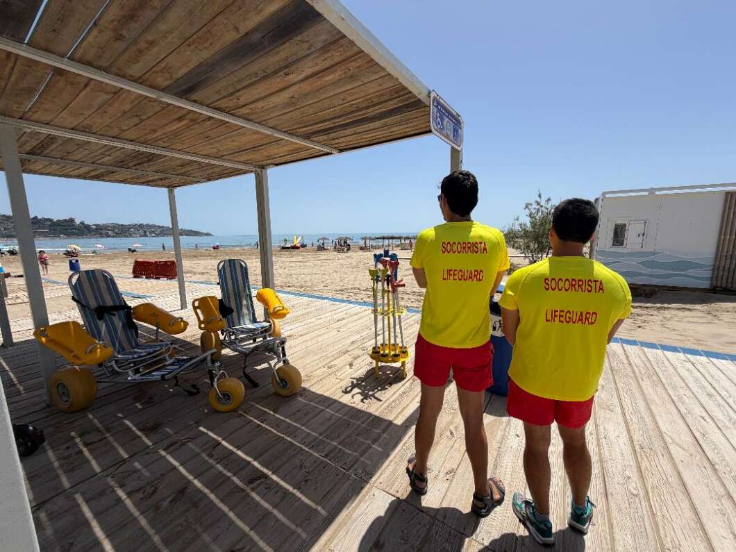 Two lifeguards in yellow shirts and red shorts stand on a wooden boardwalk at a sunny beach, with rescue gear nearby and the sea in the background.