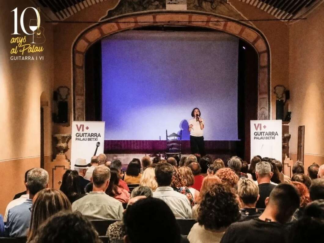 Speaker stands on stage with microphone in a concert hall, addressing a seated audience; banners read VI Guitarra Palau Betxi.