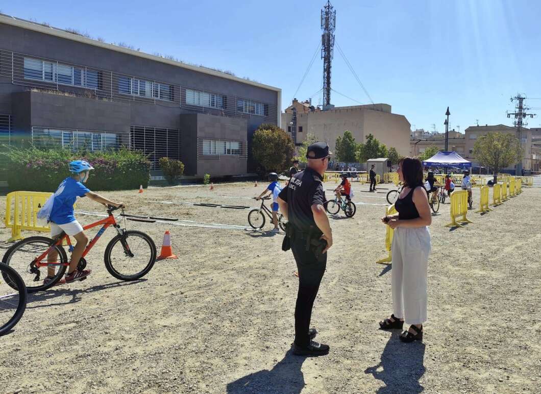 Children and adults participate in a sunny bike safety event with cones and barriers in a parking lot area under a clear sky.