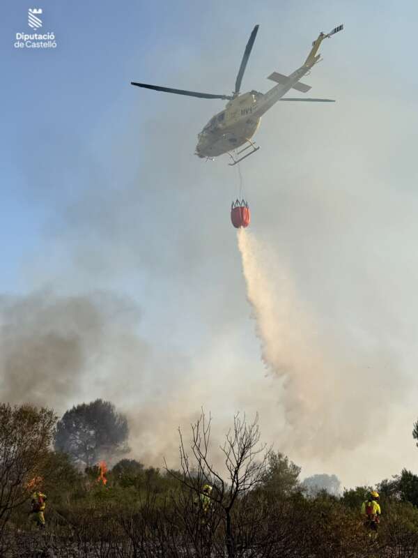 Helicopter releasing a red water bucket over a smoky brush fire, firefighters visible on the ground aiding the blaze.