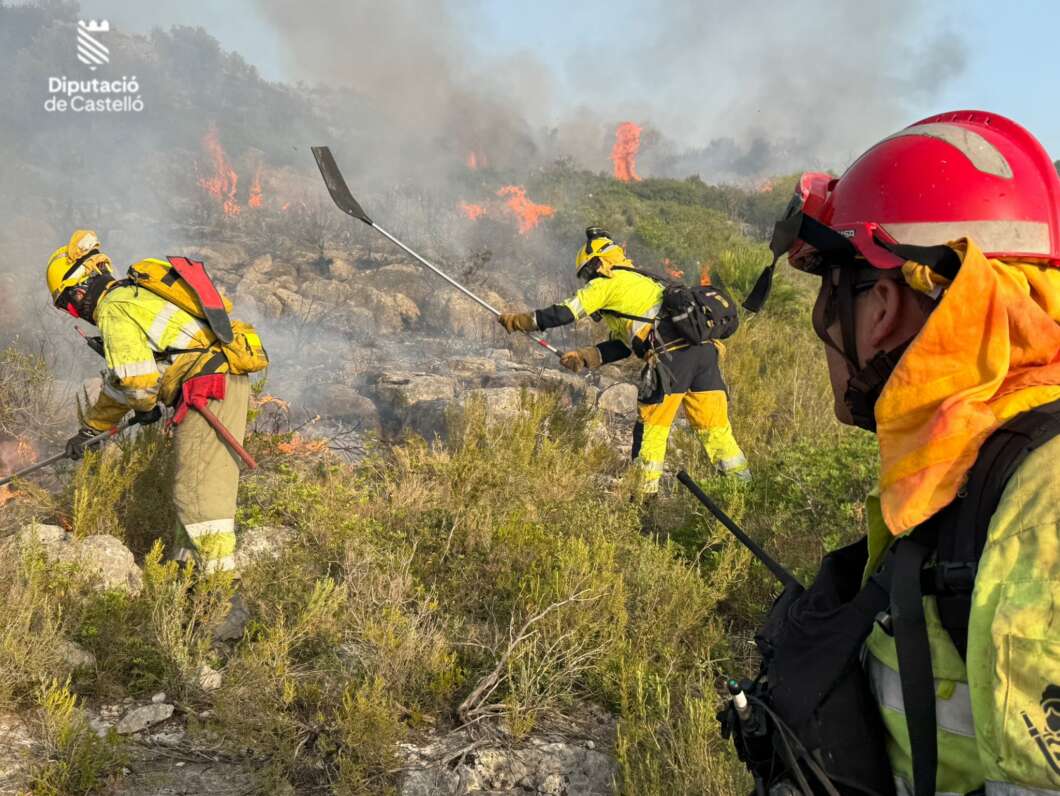 Firefighters in yellow gear and red helmets fight a wildfire on a rocky hillside with flames and smoke, logo in corner.