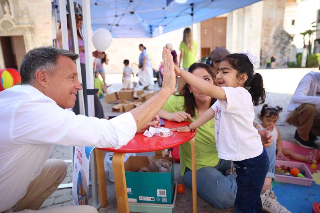 A man and a young girl high-five across a red table at an outdoor community event under a blue canopy, smiling and playing with others nearby.
