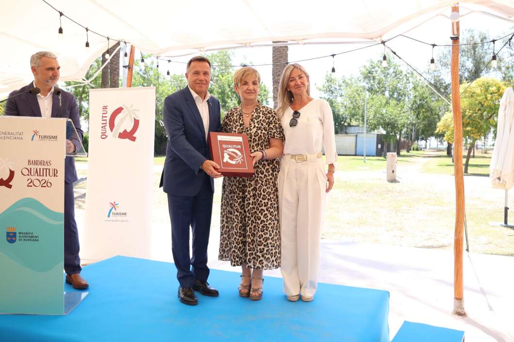 Three adults on a blue outdoor stage during an awards ceremony; a man in a navy suit presents a red plaque to a woman in a leopard-print dress, with another woman in white beside them.