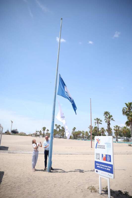 Two people stand at the base of a tall flagpole on a sunny beach as a blue flag waves overhead; palm trees and a sign are in the background.