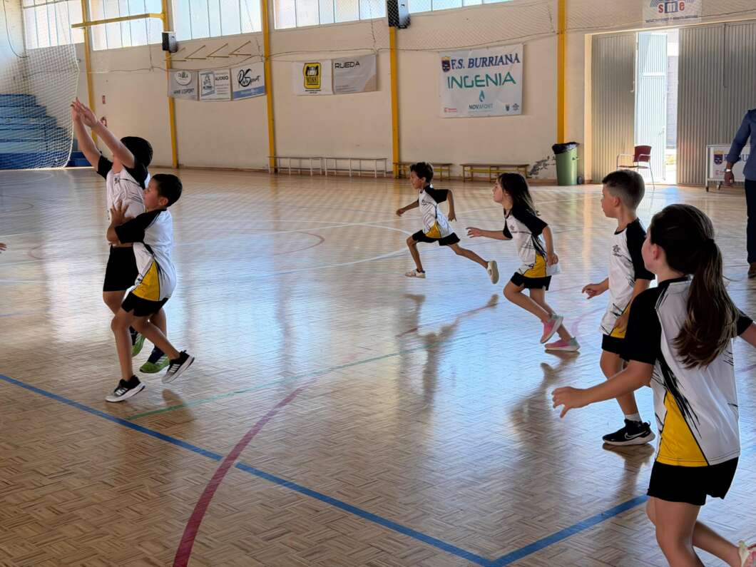 Group of children in matching white, black, and yellow uniforms running and reaching up in an indoor gym, with banners on the wall and a bright, sunlit wooden floor.