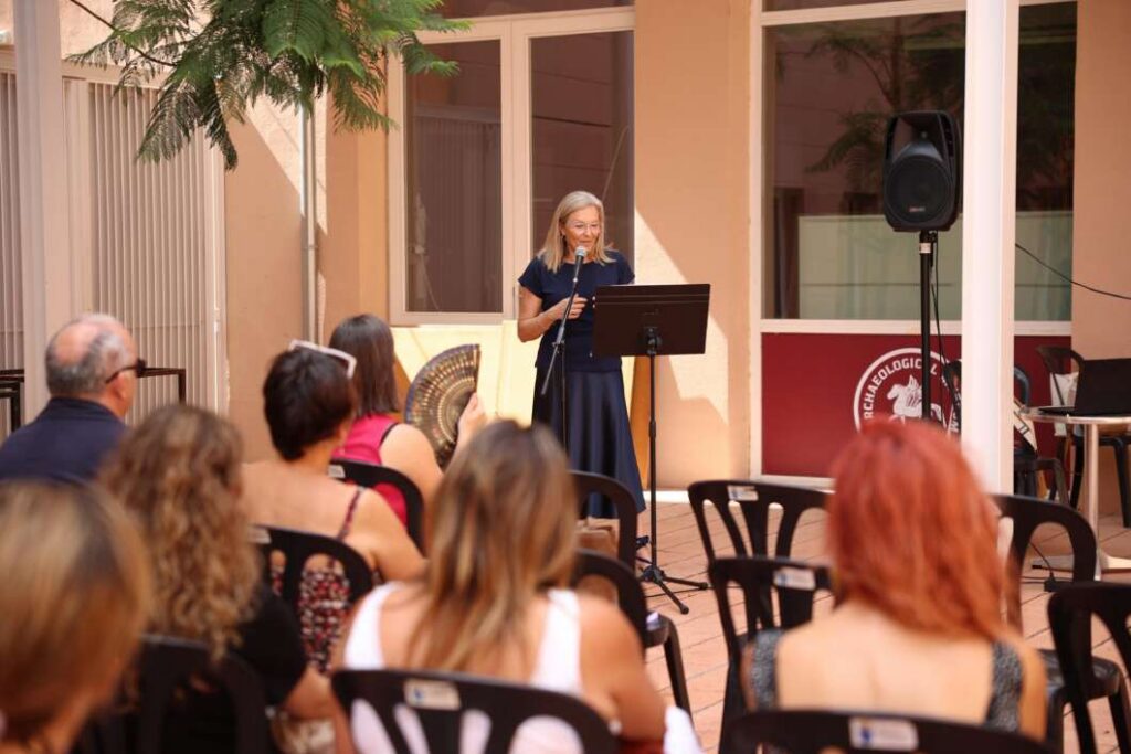 Woman in a navy dress speaks into a microphone at a stand on an outdoor patio, addressing seated attendees.