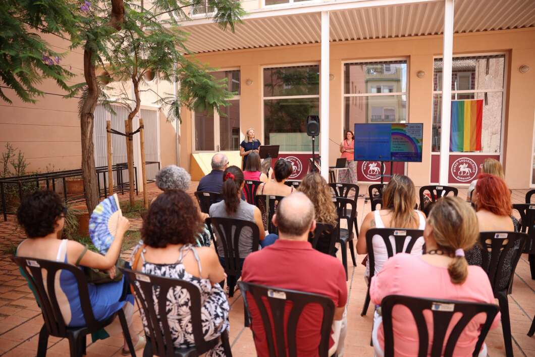 Outdoor courtyard presentation: two speakers on a small stage with a screen, audience seated on plastic chairs under a shaded canopy, rainbow flag visible.