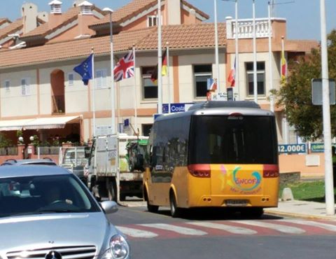 Yellow tour bus driving down a sunny street past a row of peach-colored buildings with flagpoles and flags outside a town center.
