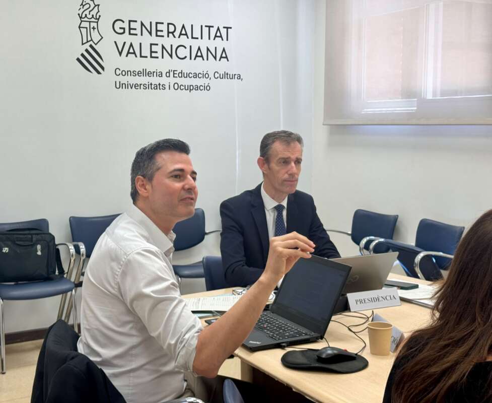 Two men in a Valencia government meeting with laptops; a 'Presidència' sign and the Generalitat Valenciana logo in the background.