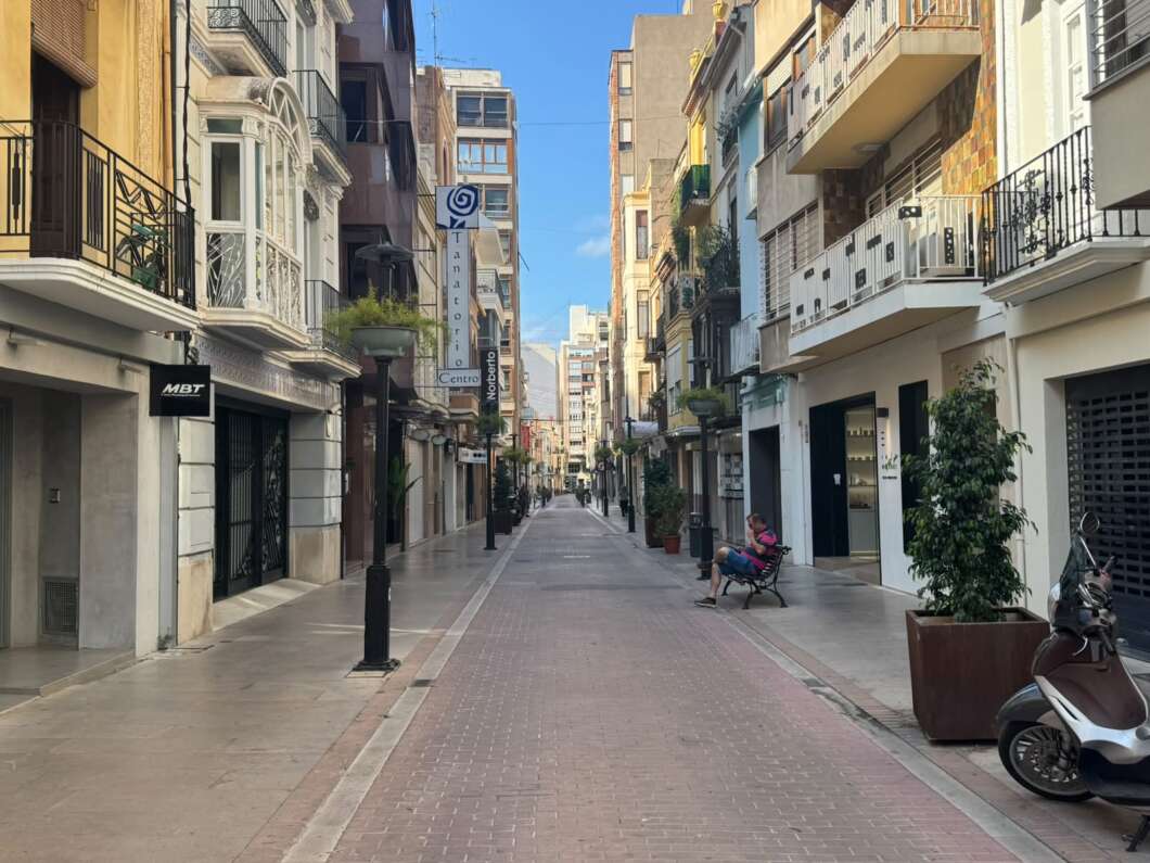 Sunlit pedestrian street with shops and balconies on both sides, a person sitting on a bench to the right and a scooter nearby.
