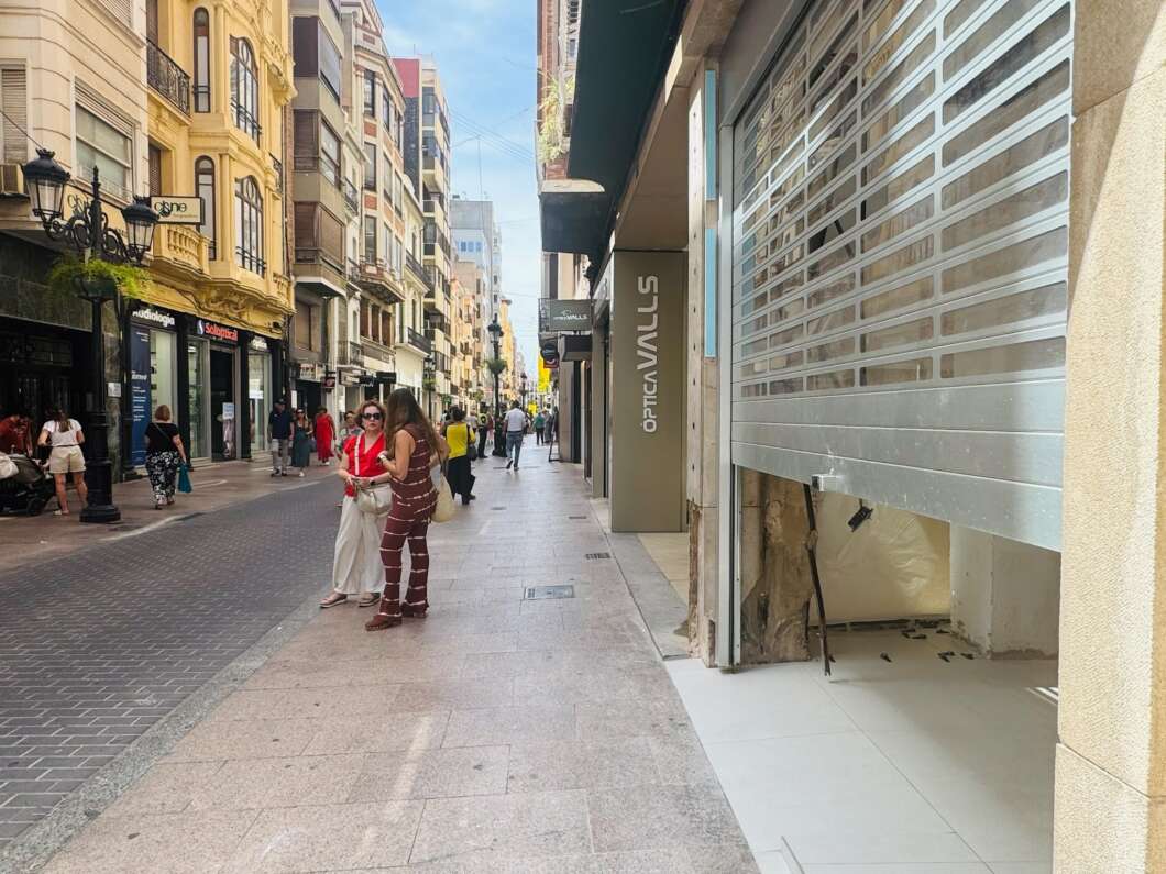 Busy pedestrian street with shoppers and colorful historic buildings on both sides; a storefront with a metal shutter on the right appears closed.