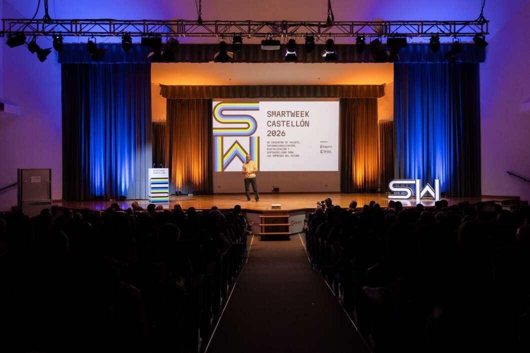 Presenter on stage delivering a talk at a conference; large screen reads 'SMARTWEEK CASTELLÓN 2026' with colorful graphic, blue/orange stage lighting, and an audience in a dark auditorium.