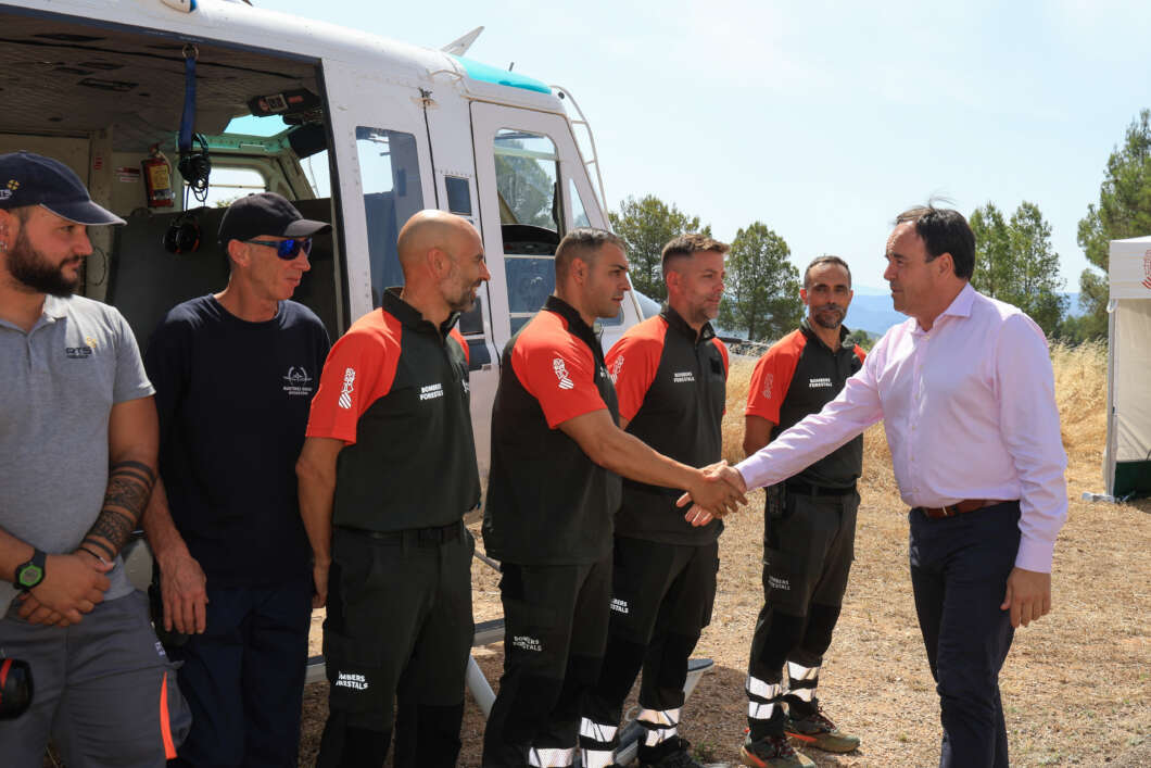 Man in a pink shirt shakes hands with a line of firefighters in black and orange uniforms beside a helicopter outdoors.