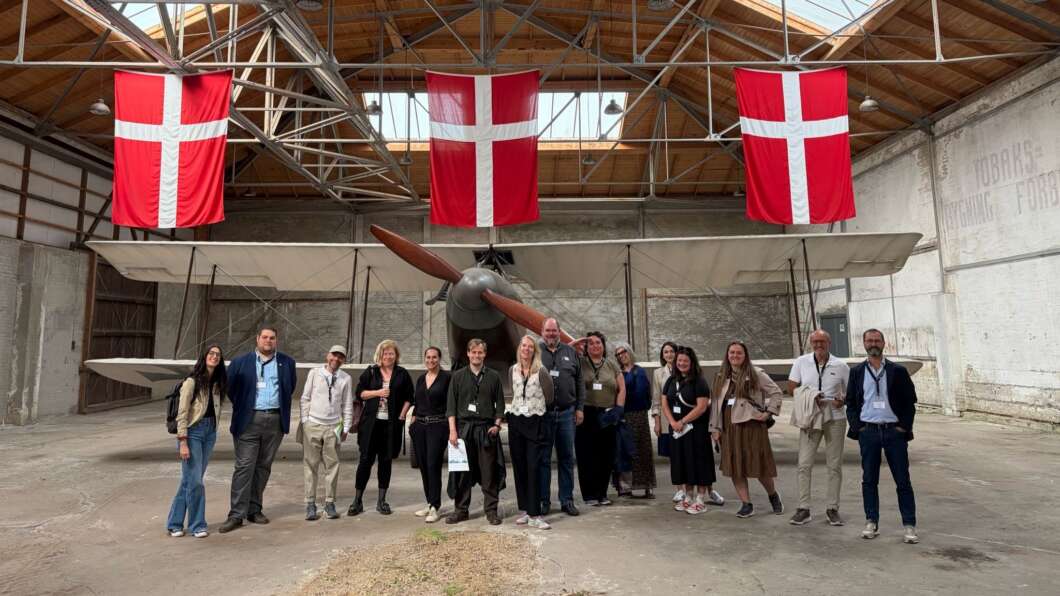 Group of people posing in a hangar with a vintage propeller airplane and three Danish flags overhead, likely on a guided tour or event.