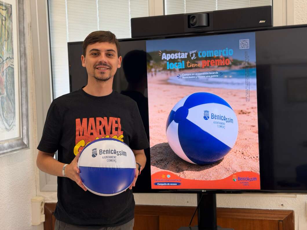 Man in a black t‑shirt holding a blue and white Benicàssim volleyball, standing beside a beach promo poster on a screen.