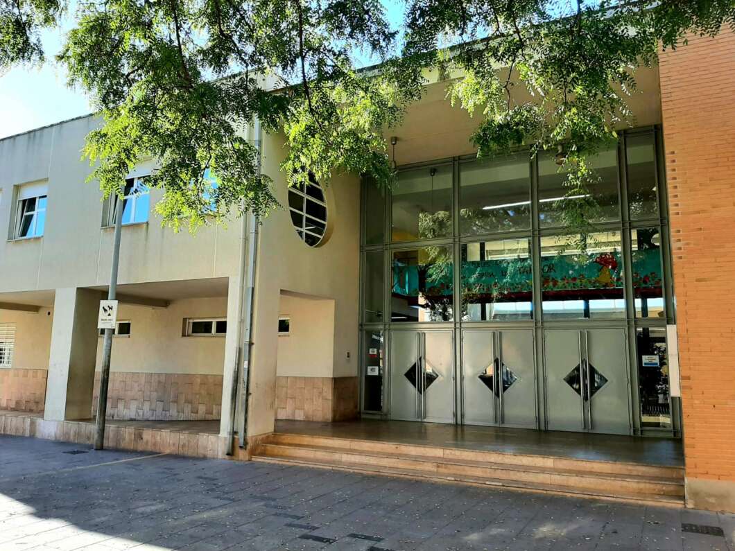 Modern building entrance with large glass doors, diamond windows, and a brick wall to the right, shaded by overhanging trees.