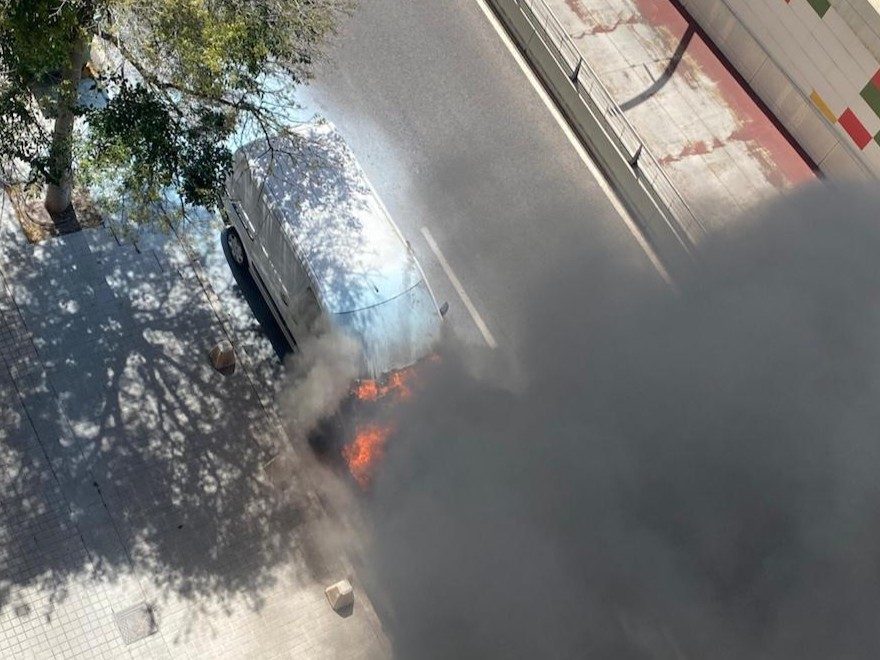 A gray SUV is on fire on a city street, with bright flames and a large plume of dark smoke rising from the engine area near the curb.