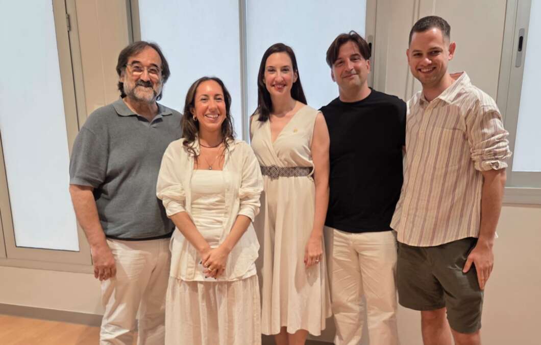 Five adults pose together indoors, smiling for the camera, dressed in light-colored outfits.