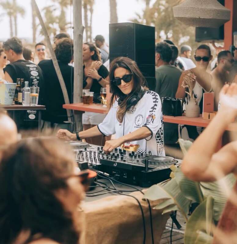 Female DJ wearing sunglasses and a white shirt mixes music at an outdoor party with a cheering crowd behind her.