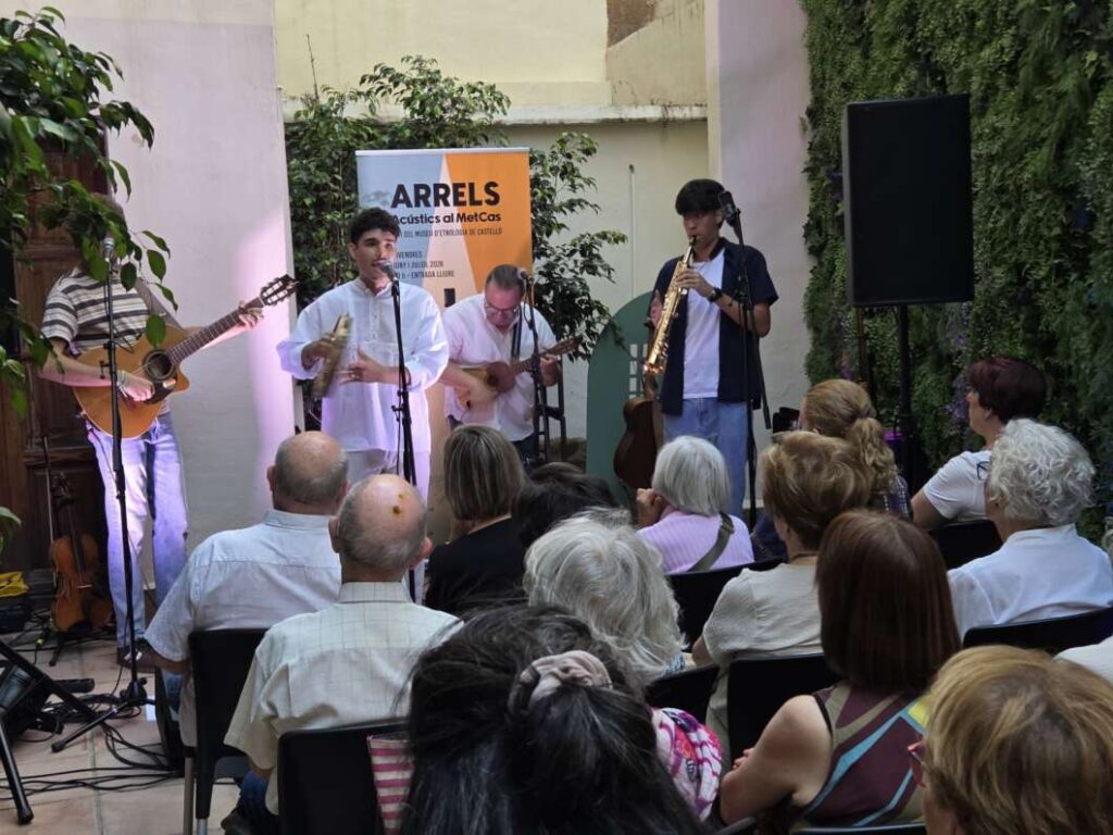 Five musicians perform a small outdoor concert in a courtyard, playing guitar, saxophone, and wind instruments while the audience watches.